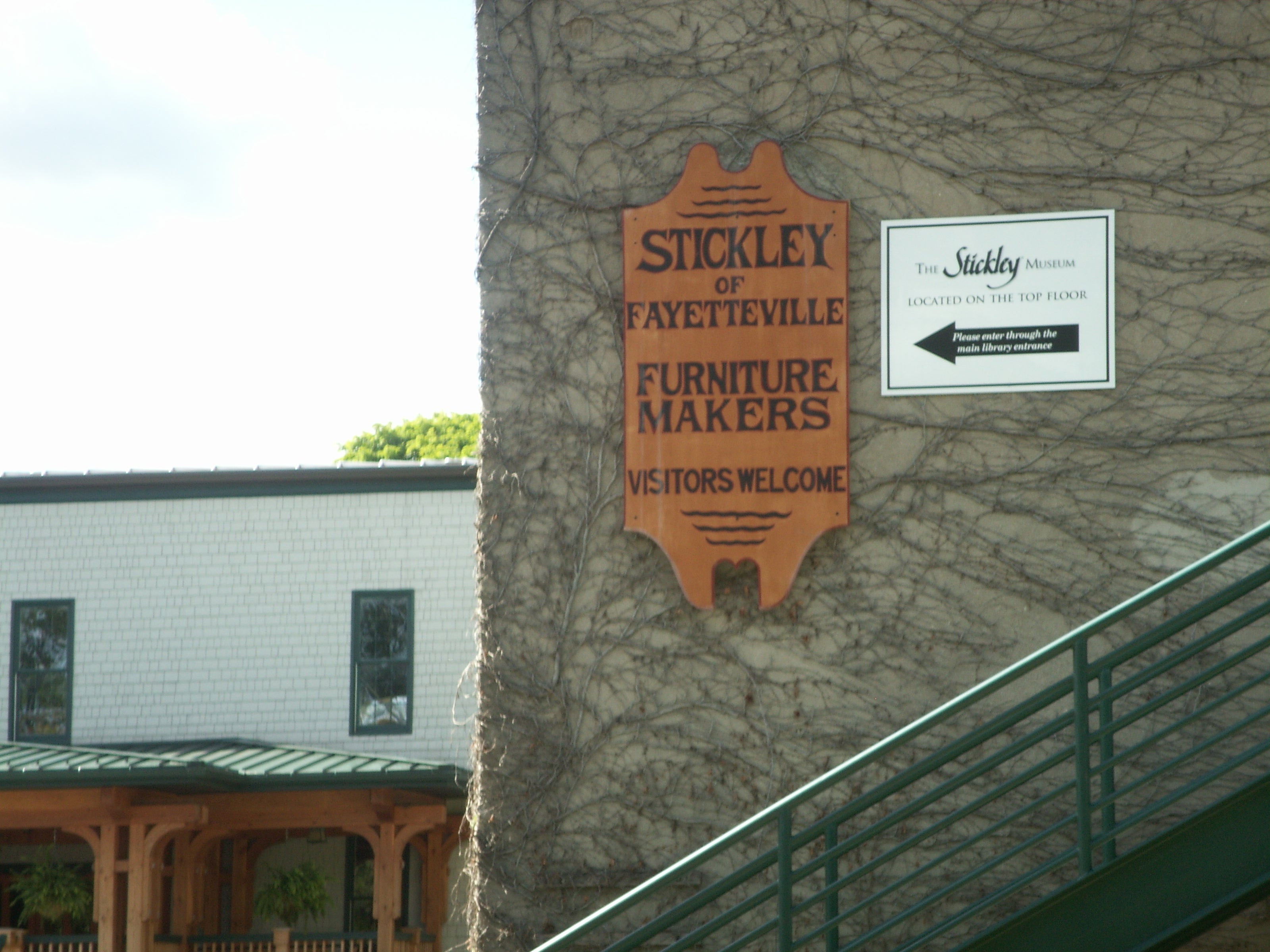 Two signs mounted on a stone wall. One is a wooden plaque reading "Stickley of Fayetteville Furniture Makers – Visitors Welcome." The other is a printed museum sign with directional instructions. A staircase runs diagonally in the foreground.